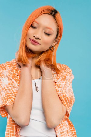 summertime happiness, radiant smile, young and charming asian woman with colored red hair posing with closed eyes and in orange shirt on blue background, generation z