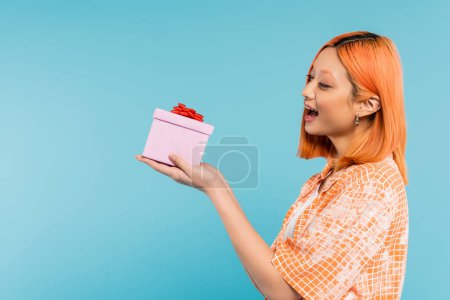 Photo for Excitement and happiness, delighted young asian woman with colored red hair and open mouth holding festive present on blue background, stylish orange shirt, youthful fashion, side view - Royalty Free Image