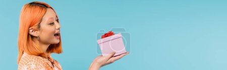 Photo for Pleasant surprise, excitement, delighted asian woman with dyed red hair and open mouth looking at gift box on blue background, happy summer, vibrant lifestyle, generation z, banner - Royalty Free Image