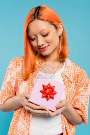Glück, entzückte Asiatin im trendigen orangefarbenen Hemd mit weißer Geschenkbox mit roter Schleife auf blauem Hintergrund, gefärbtes rotes Haar, zufriedenes Lächeln, sommerliche Stimmung, Generation z