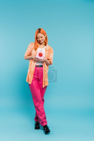 volle Länge der jungen und fröhlichen asiatischen Frau mit glücklichem Lächeln, in trendigen lässigen Kleidern stehend mit Geschenkbox auf blauem Hintergrund, gefärbte rote Haare, orangefarbenes Hemd, rosa Hose, moderne Mode