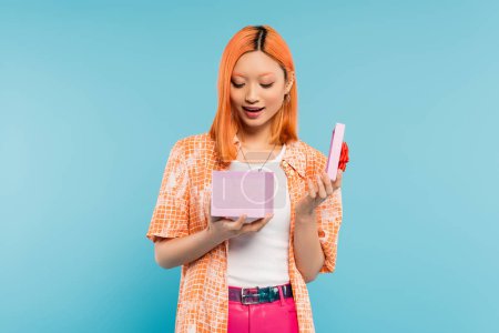 positive emotion, young, amazed and delighted asian woman with dyed red hair, in orange shirt opening gift box with festive present on blue background, surprise, joy, happiness