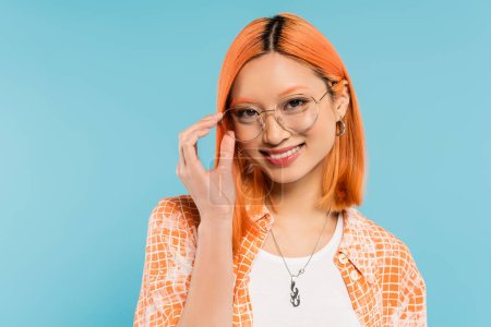 sourire radian, émotion positive, heureuse femme asiatique aux cheveux rouges teints, en chemise orange ajustant les lunettes avec cadre élégant et regardant la caméra sur fond bleu, mode jeune