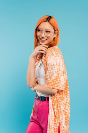 happy face, positive emotion, young and cheerful asian woman holding hand near chin and looking away on blue background, stylish eyeglasses, orange shirt, tattoo, summer style fashion