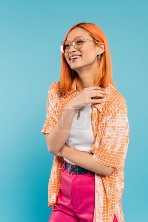 Photo for Summer and happiness, overjoyed asian woman in fashionable eyeglasses and orange shirt smiling and looking away on blue background, vibrant personality, generation z lifestyle - Royalty Free Image