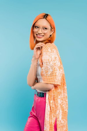 positive emotion, joyful and young asian woman with radiant smile standing and looking away on blue background, stylish eyeglasses, orange shirt, modern fashion, generation z