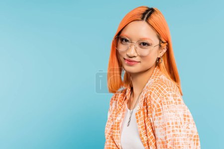 Jugendlichkeit und Glück, hübsche Asiatin mit gefärbten roten Haaren, in modischer Brille und orangefarbenem Hemd lächelnd in die Kamera auf blauem Hintergrund, Generation Z, Sommerstimmung