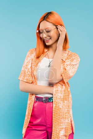positive emotion, young and cheerful asian woman adjusting colored red hair and smiling on blue background, trendy eyeglasses, orange shirt, vibrant individuality, summer fashion