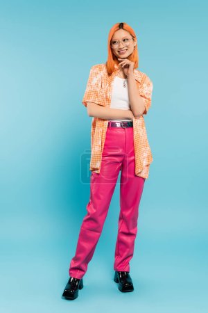 Foto de Radian sonrisa, alegre verano, longitud completa de la joven mujer asiática con la mano cerca de la barbilla mirando hacia otro lado sobre fondo azul, pelo rojo teñido, gafas de moda, camisa naranja, pantalones de color rosa, moda juvenil - Imagen libre de derechos