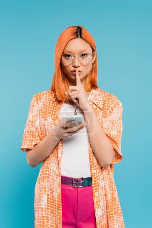 secret, silencieux s'il vous plaît, jeune femme asiatique tenant smartphone, regardant la caméra et montrant signe de silence sur fond bleu, cheveux rouges teints, lunettes à la mode, chemise orange, mode d'été