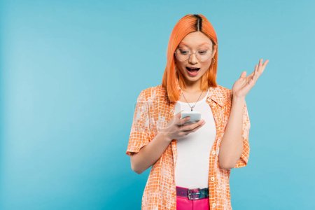 asombro, mujer asiática joven con el pelo rojo de color y la boca abierta mirando el teléfono móvil y el gesto sobre fondo azul, gafas de moda, camisa naranja, moda juvenil, estilo de vida digital