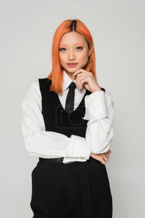 business casual clothes, portrait of captivating asian woman with colored red hair, in white shirt, black tie and vest holding hand near face and looking at camera on grey background