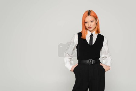 femme asiatique auto-assurée avec des cheveux roux colorés posant avec les mains dans les poches en chemise blanche, cravate noire, gilet et pantalon lorsque vous regardez la caméra sur fond gris, business casual fashion