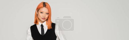 Téléchargez les photos : Portrait de femme asiatique séduisante et à la mode regardant la caméra sur fond gris, cheveux rouges colorés, chemise blanche, cravate noire et gilet, style décontracté d'affaires, génération z, bannière - en image libre de droit