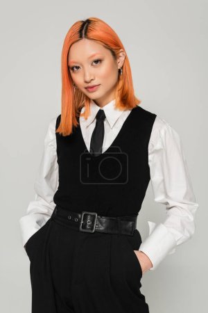 portrait of charming and young asian woman holding hands in pockets and looking at camera on grey background, dyed red hair, white shirt, black tie, vest and pants, business fashion photography