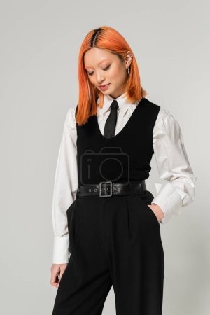 femme asiatique élégante et séduisante avec des cheveux rouges teints, en chemise blanche, cravate noire, gilet et pantalon posant avec la main dans la poche sur fond gris, génération z style de vie, mode décontractée d'affaires