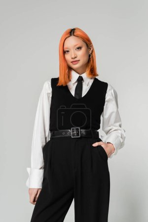 attractive asian woman with expressive gaze holding hand in pocket and looking at camera on grey background, dyed red hair, white shirt, black tie, vest and pants, business fashion, youth culture