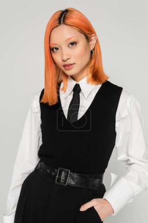 beautiful and trendy asian woman with dyed red hair, in white shirt, black tie, vest and pants holding hand in pocket and looking at camera on grey background, business casual, youth culture