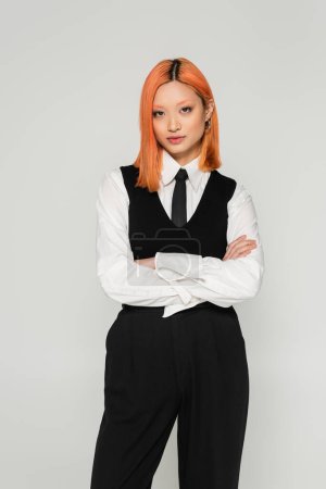youthful and self-assured asian woman standing with crossed arms and looking at camera on grey background, colored red hair, white shirt, black tie and pants, youthful fashion, business casual 