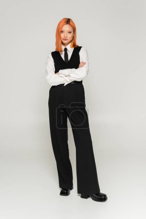 modern and confident asian woman in trendy casual clothes standing with folded arms on grey background, colored red hair, white shirt, black tie, vest and pants, business casual fashion