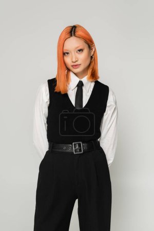 modern and pretty asian woman with colored red hair standing with hands behind back while posing in white shirt, black tie, vest and pants on grey background, business casual, youthful style