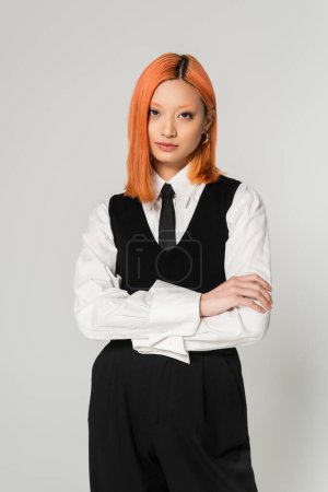 portrait of attractive and young asian woman in white shirt, black tie, vest and pants standing with folded arms on grey background, dyed red hair, confident gaze, looking at camera, business casual