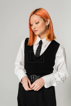 mujer asiática encantadora y positiva con el pelo rojo de color de pie y sonriendo sobre fondo gris, camisa blanca, corbata y chaleco negro, moda casual de negocios, generación z, estilo de vida moderno