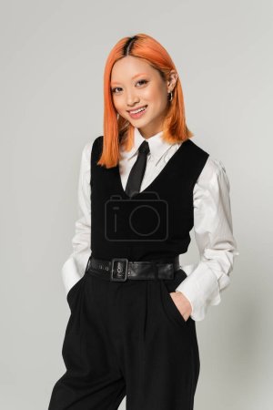cheerful emotion, appealing and stylish asian woman with radiant smile holding hands in pockets and looking at camera on grey background, black and white business casual clothes, generation z