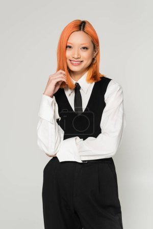 happy emotion, young and joyful asian woman with dyed red hair looking at camera on grey background, white shirt, black tie, vest and pants, business casual fashion photography, modern lifestyle