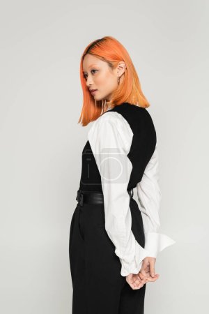 Photo for Side view of young and charming asian woman with colored red hair, in white shirt and black vest standing with hands behind back and looking away on grey background, business casual style - Royalty Free Image