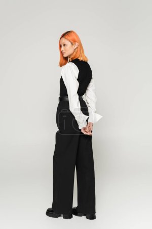 fashion photography of young asian woman with colored red hair, in white shirt, black vest and pants standing with hands behind back and looking away on grey background, business casual style