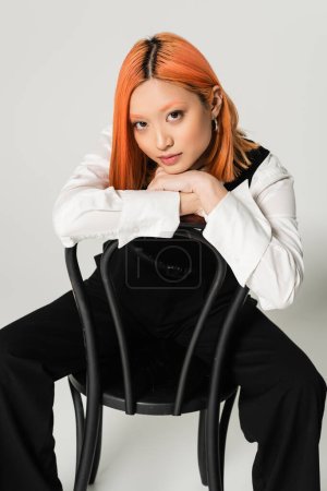 youthful and pretty asian woman with dyed red hair wearing white shirt, black vest and pants while sitting on chair and looking at camera on grey background, business fashion photography