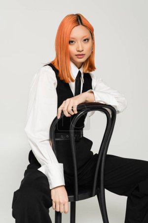 femme asiatique expressive avec les cheveux rouges teints assis sur la chaise et posant à la caméra sur fond gris, cheveux rouges colorés, vêtements noirs et blancs, photographie de mode d'affaires, génération z