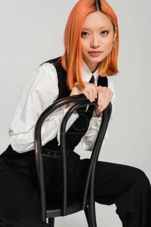 femme asiatique charismatique et à la mode avec des cheveux rouges colorés, chemise blanche, gilet noir et pantalon posant sur la chaise et regardant la caméra sur fond gris, mode d'affaires moderne
