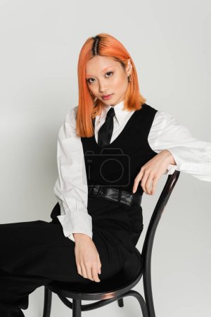 junges und bezauberndes asiatisches Model mit roten Haaren, die in die Kamera schauen und auf einem Stuhl auf grauem Hintergrund sitzen, weißes Hemd, schwarze Weste, Krawatte und Hose, Business Casual Fashion, moderner Stil