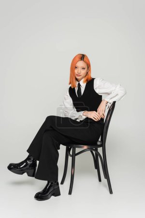 attractive and self-assured asian woman with colored red hair, in white shirt, black vest and pants sitting on chair and looking at camera on grey background, fashion shoot, full length