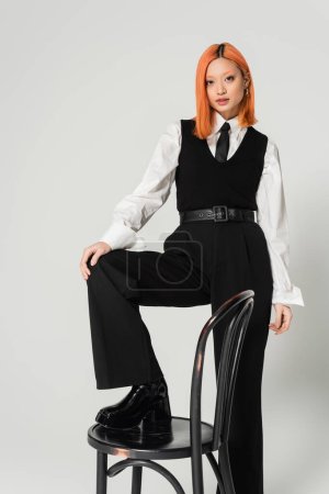 fashion shoot, attractive asian woman in white shirt, black vest, tie and pants, with colored red hair stepping on chair and looking at camera on grey background, generation z