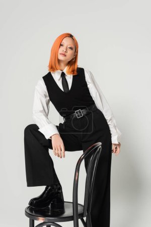 Photo for Pretty and confident asian woman in white shirt, black tie, vest and pants, with colored red hair stepping on chair and looking at camera on grey background, modern business fashion, generation z - Royalty Free Image