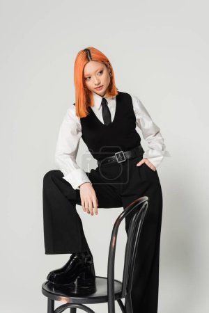 young and confident asian woman holding hand in pocket and looking away while posing with chair on grey background, dyed red hair, black and white business casual clothes, fashion shoot