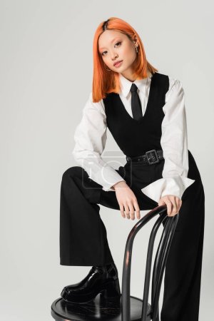 charmante und trendige asiatische Frau, die in die Kamera schaut, wenn sie auf einem Stuhl auf grauem Hintergrund posiert, gefärbte rote Haare, schwarz-weiße Business-Freizeitkleidung, Lebensstil der Generation Z