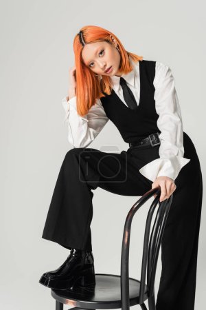 young and attractive asian woman with red colored red hair, in white shirt, black tie, vest and pants posing with chair on grey background and looking at camera, modern business fashion