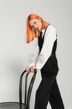 casual business fashion photography, red haired asian woman in white shirt, black tie, vest and pants looking at camera near chair on grey background, youth culture, generation z