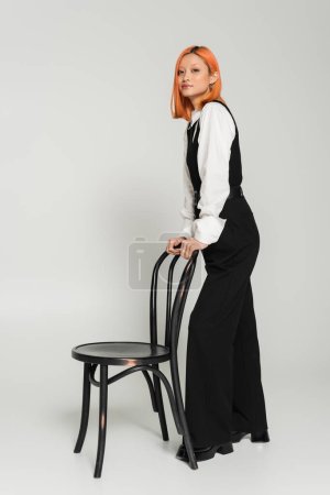 expressive and red haired asian woman standing near chair and looking at camera on grey background, stylish black and white clothes, business casual, modern lifestyle, full length