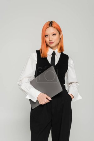 attractive asian woman with colored red hair, in black and white clothes holding laptop and looking at camera on grey background, white shirt, black vest, freelance lifestyle, generation z