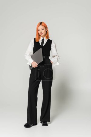 Photo for Positive and confident freelancer, young asian woman with red colored hair standing with hand in pocket and laptop on grey background, business casual, white shirt, black vest and pants - Royalty Free Image