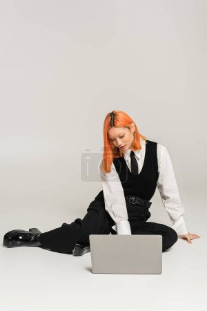 business casual fashion, young asian woman with red dyed hair working on laptop while sitting on grey background, white vest, black vest and pants, freelance lifestyle, generation z