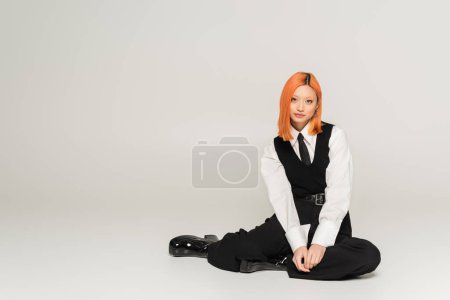 full length of confident and successful asian woman sitting and looking at camera on grey background, business casual, black vest, tie and pants, white shirt, colored red hair, youthful style