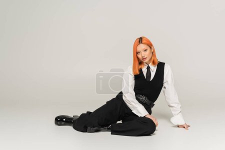 full length of stylish and confident asian woman with dyed red hair sitting and looking at camera on grey background, business casual, black vest and pants, white shirt, modern fashion, gen z