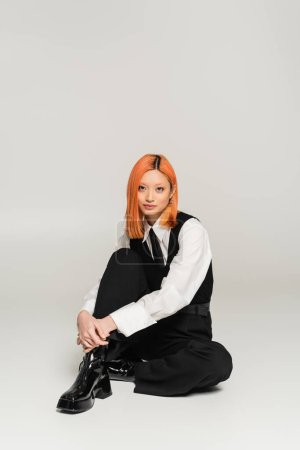 charming and self-assured asian woman with colored red hair looking at camera when sitting on grey background, white shirt, black vest and pants, business casual fashion, modern lifestyle
