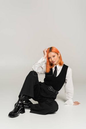 young asian woman with colored red hair, in black and white clothes sitting and looking at camera on grey background, white shirt, black pants and vest, business casual fashion, full length